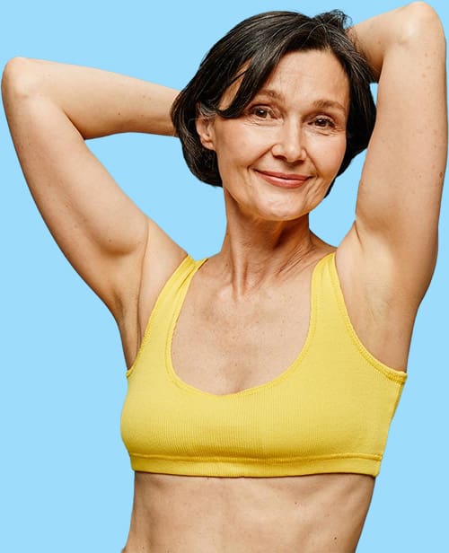 Ältere Frau im gelben Sport-BH zeigt ihre haarfreien Achseln vor blauem Hintergrund.